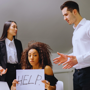 Woman holding sign that says help with two people standing over her and gesturing while frowning